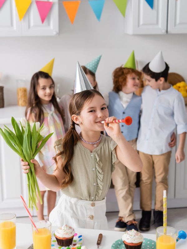 cheerful girl holding tulips and blowing party horn near friends during birthday party at home